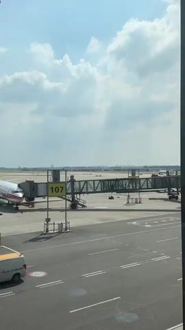 The video shows an airport tarmac. A jet bridge, labeled with the number 107, is connected to a gate. In the distance, a passenger airplane is taxiing on the runway. The sky is partly cloudy.
