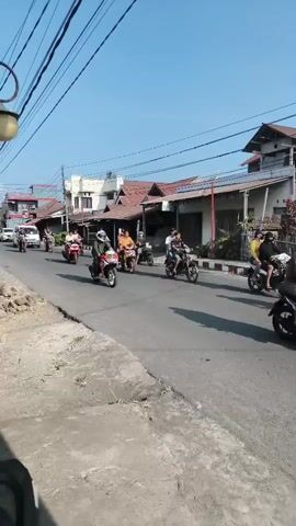 A group of motorcyclists are riding down a street. The motorcyclists are riding in single file and are all wearing helmets. The street is lined with buildings on either side. The sky is clear and blue.