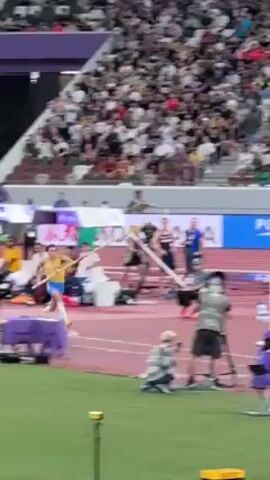 A pole vaulter in a yellow and blue uniform clears the bar and lands safely on the mat. There is a large crowd in the background and a TV camera crew and officials on the infield of the track and field stadium. There are advertising banners of sponsors in the background of the shot.