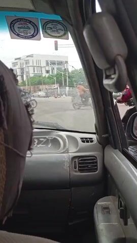 The video, filmed from inside a vehicle, shows a street intersection. Several motorcycles and cars are visible, along with a traffic light that is red. Buildings and trees line the street. Two stickers are affixed to the windshield, one with a green outline and text, and another that appears to be a circular logo. The dashboard of the vehicle is visible, showing the "BZT" label and air vents. A headrest is also visible in the foreground. The lighting suggests it is daytime.