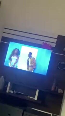 A television shows a music video of a man and woman singing and moving. The woman is wearing a light blue dress, and the man is wearing a khaki outfit. The background of the video is light blue and white. The television is mounted on a dark wood console. A plastic bottle, a white container, and dark-colored objects are on or near the console. The wall behind the television is white with dark wood trim.