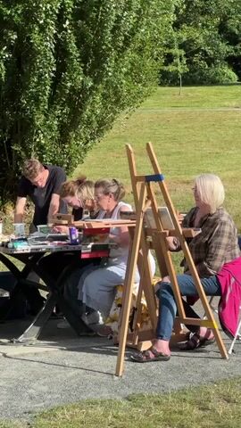 A group of people are gathered around a table outside, engaged in an art class. A man in a black t-shirt stands at the left side of the table, appearing to instruct or assist the others. Three women are seated at the table, each with art supplies in front of them. The woman in the center is wearing a light blue dress and is actively painting. To her right, another woman in a plaid shirt sits at an easel, also painting. The environment is a park-like setting with trees and grass visible in the background, bathed in sunlight.