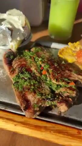 Here's a description of the video: 
The video shows a close-up of a meal on a wooden table. The main dish consists of a grilled steak covered in green herbs and red peppers. Next to the steak are plantain patties topped with a red sauce and a baked potato wrapped in foil with white cream or butter on top. A green juice is in a glass with a white straw in the background. The food is presented on a metal tray. Steam is rising from the steak. There are other out-of-focus items in the background, including a purse and a red object. The camera is relatively static. The lighting appears to be natural, possibly from sunlight.