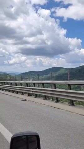 The media file shows a view from a vehicle driving on a highway in a mountainous region. The road has guardrails along the side, and tire tracks are visible on the asphalt. In the background, lush green hills and mountains are covered with trees. The sky is partly cloudy with a mix of bright blue sky and white, puffy clouds. Sunlight suggests it is daytime. In the distance, a large bridge structure can be seen crossing a valley. The overall scene conveys a sense of travel and the scenic beauty of a natural landscape. The specific location is Vrachesh, Bulgaria.