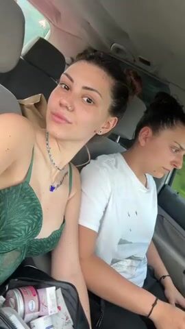 Two women are filmed inside a car during the daytime in Stolnik, Bulgaria. The woman on the left has dark hair styled in two buns and is wearing a green lace top, a necklace with a blue pendant, and a nose ring. She smiles directly at the camera, then turns and gives a quick kiss on the cheek to the woman seated next to her on the right. This second woman also has dark hair in a bun and is wearing a white t-shirt featuring a graphic print of mountains, water, and birds, along with some text that is partially visible, possibly reading "KEEP THE MOST CALM". She appears more subdued, looking down or away from the camera, but reacts to the kiss. In the foreground, a bag containing several small containers with visible labels, including "BALÉA" and "SKIN," rests on the seat. The bright light suggests it is sunny outside, and the car appears to be in motion.