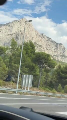 The video shows a view from inside a vehicle traveling on a road. On the right side of the road, there is a guardrail, followed by a dense green forest. Behind the forest, a large, rocky mountain dominates the background, with a partly cloudy blue sky above. A tall lamppost stands near the road. Further down the road, there are signs and a billboard with images. The road has lane markings, and other vehicles are visible in the distance. The sun appears to be shining.