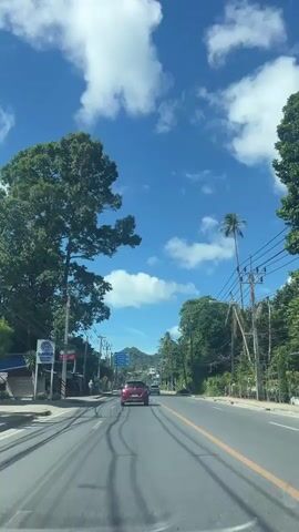 A first-person perspective video shows a car driving on a road with a bright blue sky and white clouds. Lush green trees line both sides of the road, and there are power lines running overhead. Several other vehicles, including cars and a scooter, are visible on the road. On the left side of the road, there is a sidewalk with a few people walking. There are also some buildings and signs visible. The road appears to be in a tropical or subtropical area due to the vegetation and palm trees.