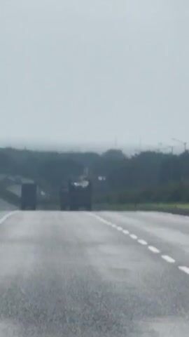 A video captures a highway scene with two trucks driving away from the camera. The road is wet, suggesting recent rain. On the left side of the highway, a motorcycle and rider are visible. The sky is overcast, and the distant landscape is blurred, creating a hazy atmosphere. Streetlights line the left side of the road. The overall impression is one of a quiet, possibly damp, day on a highway.