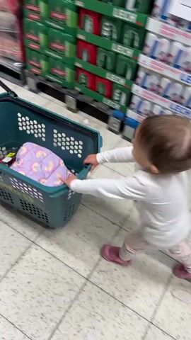 A toddler is pushing a small, teal-colored shopping basket down a long aisle in a supermarket. The basket has a metal handle. The child is wearing a white long-sleeved shirt, patterned light-pink pants, and pink shoes. Her hair is in a small ponytail held by a pink hair tie. She is facing away from the camera. The supermarket aisle has shelves filled with various products on both sides, including drinks, food, and other grocery items. The floor is tiled. The video follows the child as she pushes the basket along the aisle.