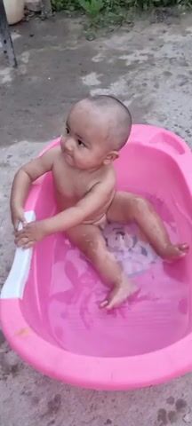 A young baby, likely a toddler, is seated in a pink baby bathtub filled with clear water. The baby is bald, with light skin, and is looking to the left of the frame. They are naked and appear to be enjoying their bath, as indicated by their relaxed posture and the splashing of water. A cartoon character, possibly Mickey Mouse, is visible on a mat at the bottom of the bathtub. The bathtub is placed on a rough, gray surface, possibly concrete or dirt, suggesting an outdoor setting. The lighting appears to be natural, indicating daytime, though the specific time of day and weather are not discernible from the image. There is no visible text in the media file. The context provided places this scene in Jomi, Tajikistan.
