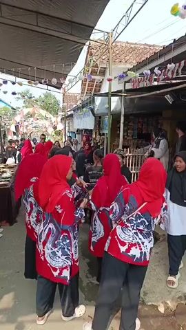 A group of young people wearing red hijabs and red shirts with a white and blue dragon design are standing together. They are wearing black pants. One person in the back is wearing a black hijab. In the background, there are market stalls with various items for sale, decorated with colorful flags and paper lanterns. There are other people in the background, some of whom appear to be vendors or shoppers. The scene appears to be an outdoor market or festival.