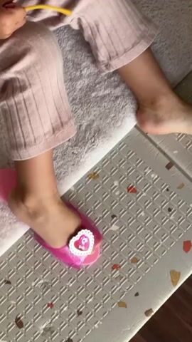 The video shows a close-up of a child's legs and feet. The child is wearing pink, ribbed pajama pants and a pink slipper with a heart-shaped gem. The child's left foot is resting on a white, textured mat, and the right foot is on a gray mat with a textured grid pattern. The child's feet are moving slightly, and the slipper appears to be a play shoe. In the background, the child's torso and upper body are visible, along with a glimpse of their hair and yellow glasses. The scene suggests a domestic environment, possibly a playroom or bedroom.
