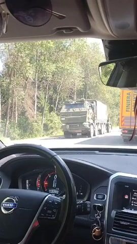 The video is filmed from the interior of a car, looking forward through the windshield. The driver's hands are visible on the steering wheel, and part of the car's dashboard is in the foreground. Outside the car, a road is visible, lined with trees. In the distance, a large, olive-green military truck is driving on the road, followed by a yellow truck. The military truck appears to be moving towards the camera, or at least approaching the car's position. The sunlight suggests it is daytime, likely in a temperate climate.