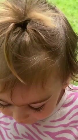 The video shows a young child with blonde hair, wearing a pink and white striped t-shirt and pink shorts, sitting on a wooden swing in a grassy yard. The child is holding onto the ropes of the swing and appears to be smiling and laughing. The camera zooms in on the child's face at one point. The background consists of green grass and foliage. The video captures the child swinging back and forth.