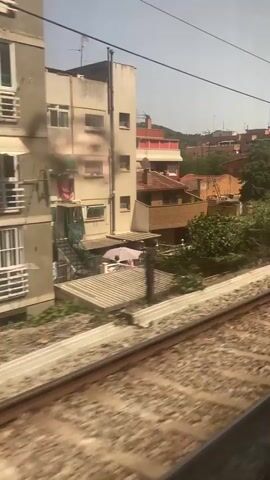 A video captures a view from a moving train looking out a window. The train tracks are in the foreground, blurred due to motion. Beyond the tracks, a row of residential buildings with terracotta roofs are visible, interspersed with lush green trees and foliage. Some buildings have balconies and windows, and one building has graffiti on its lower section. Hills covered in vegetation rise in the background under a hazy sky. The camera pans slowly, showing different parts of the landscape as the train travels.
