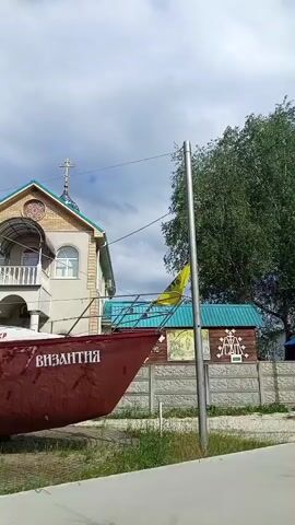 The video shows a red boat with the word "BYZANTIA" written on its side, parked in front of a building that resembles a church. The church has a green roof with a cross on top and a balcony on the second floor. A yellow flag is visible on the boat, attached to a pole. The scene appears to be outdoors, with trees and a cloudy sky in the background. The boat is resting on some wooden supports.
