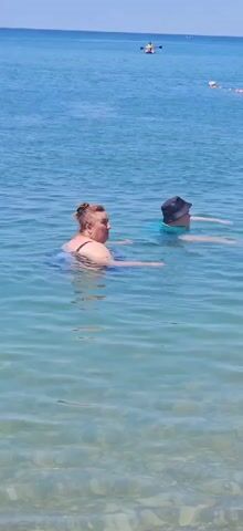Two people are swimming in clear, blue water. The person in the foreground is a woman with red hair pulled up in a bun, wearing a dark swimsuit. The person behind her is wearing a blue shirt and a dark hat. They are both looking to the right. The water is shallow near the shore, with pebbles visible on the bottom.