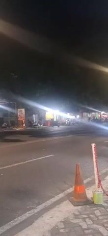 A dimly lit, outdoor scene at night. In the foreground, a paved sidewalk is visible, with a traffic cone and two stanchions connected by red chains. The road in front of the sidewalk is dark with some faint white lines indicating lanes. Several vehicles, including what appear to be motorcycles or scooters, are present on the road and are illuminated by headlights and streetlights. In the background, there are multiple stalls or shops with bright, artificial lighting, suggesting a commercial area, possibly a night market or food court. Trees line the edge of the road and the background. A large red sign with white text is visible in the upper right corner of the frame, partially obscured by glare. The overall atmosphere is busy and active, despite the low light conditions.