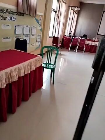 The video shows a room that appears to be a meeting or presentation space. There are several tables covered with red tablecloths, some arranged in rows and others in a circular formation. Many red chairs are positioned around the tables. A large projection screen is visible on one wall, and a whiteboard or bulletin board with charts and papers is on another wall. The room has tiled flooring and neutral-colored walls. There are windows with blinds, and what looks like an air conditioning unit. The lighting suggests it is daytime. No people are visible in the video.