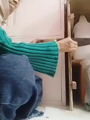 A person wearing a headscarf, a green sweater, and blue jeans is kneeling on the floor next to a wooden cabinet. The person is reaching into the cabinet and appears to be moving something. The cabinet has shelves, and there are white containers on the shelves. The environment appears to be indoors, with a white wall in the background.