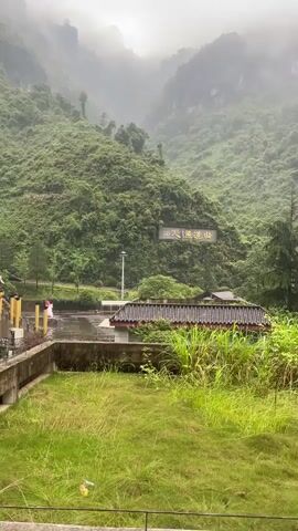 The video shows a view of a traditional Chinese-style building complex against a backdrop of misty green mountains under an overcast sky. The building has a dark-colored roof with golden ornamental details along the eaves. The mountains are densely covered in green vegetation and partly obscured by the fog. The foreground includes some greenery and a structure, partially hidden, which seems to lead to the building. The atmosphere is calm and somewhat muted due to the weather.