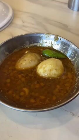 The video shows a close-up shot of a bowl of food. The bowl is made of metal and contains a brown, gravy-like sauce with chickpeas and two boiled eggs. There is also a green chili pepper in the bowl. Next to the bowl, on a white plate, is a piece of flatbread with a dimpled surface. The food is placed on a marble-patterned table. In the background, there are stacks of white plates and wooden chairs. The lighting is bright and natural, suggesting it might be daytime.