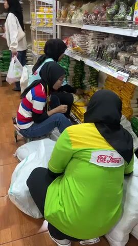 Three individuals wearing headscarves are in an aisle of a store, surrounded by shelves stocked with various products. The person in the foreground, facing away from the camera, wears a bright green shirt with a yellow panel on the shoulders and a logo that reads "Teh Jawa". They are kneeling on large white sacks. In the middle ground, a person wearing a red, white, and blue striped shirt and blue jeans is seated on a small stool, also wearing a headscarf. They appear to be examining or handling a yellow package. Behind them, another person in a teal shirt and headscarf is kneeling and reaching towards the shelves. The shelves are filled with packaged goods, including what appear to be bags of rice and other food items. The floor is tiled. The overall impression is of an inventory or stocking activity within a retail environment.