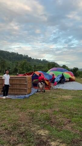 A group of people are preparing a hot air balloon for flight in an open field. The balloon, a vibrant patchwork of colors including purple, green, yellow, orange, and pink, is partially inflated and lies spread out on the ground. Several individuals in red shirts are actively working on the balloon, appearing to adjust its fabric. Adjacent to the balloon is a wicker basket, presumably the gondola, also being prepared. A man in a white shirt and black pants stands to the left of the basket, observing the process. The environment is a grassy field with a line of trees and hills in the background under a cloudy sky.