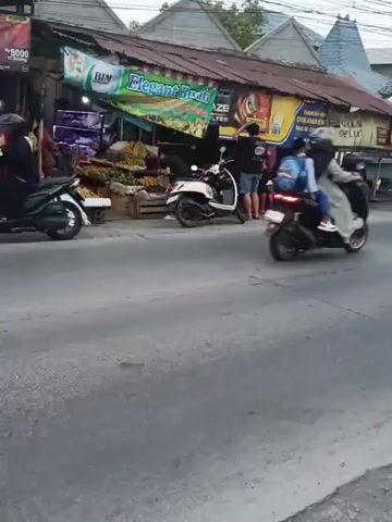A street scene with a row of market stalls on the right side of the road. The stalls have corrugated metal roofs and appear to be selling produce. There are several motorcycles parked in front of the stalls and people can be seen browsing or interacting. A white scooter is parked in front of one of the stalls. On the left side of the road, a black van is driving away from the camera. Traffic appears to be moving slowly. The weather looks overcast.