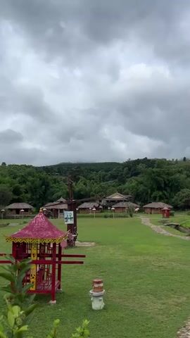 The video shows a panning shot of a village with thatched-roof buildings and green grass. In the foreground, there is a red gazebo with intricate details. The buildings are low and appear to be made of brick and wood, with decorative lanterns hanging from them. The landscape is hilly and covered with lush green trees. The sky is cloudy and overcast, suggesting a potentially rainy day. The overall atmosphere is serene and picturesque.