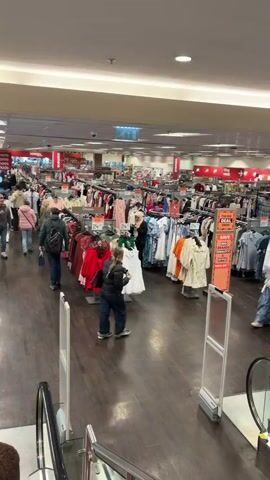 The camera pans slowly across a busy department store floor. Rows of clothing racks densely packed with various garments are lined up, with sale signs interspersed. Shoppers are browsing the merchandise, some walking individually, others in small groups. The floor is dark wood, and the overhead lighting is bright and uniform. In the foreground, the top of an escalator is visible. Signage indicates the directions to different departments, such as "Home" and "Kids." The overall impression is of a bustling retail environment.