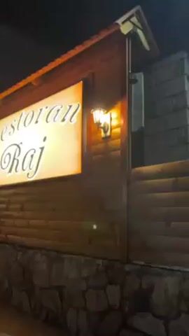 The video shows the exterior of a restaurant at night. The restaurant has a wooden facade with a stone base. A large illuminated sign reads "Restoran Raj" in cursive lettering. The sign is flanked by two lantern-style lights. A set of stone steps leads up to a wooden gate, visible in the background to the left of the sign. The sky is dark, and the overall scene is dimly lit.