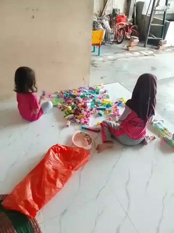 Two young girls are sitting on a tiled floor, playing with a pile of colorful building blocks. The girl on the left is wearing a pink shirt and has dark hair. She is sitting facing away from the camera. The girl on the right is wearing a pink headscarf and a pink shirt with gray pants. She is sitting facing the blocks. In the foreground, there is a large orange plastic bag and a small pink bowl. The background shows a cluttered area with furniture, a bicycle, and some brickwork, suggesting an outdoor or semi-outdoor space. The lighting is natural, indicating daytime.