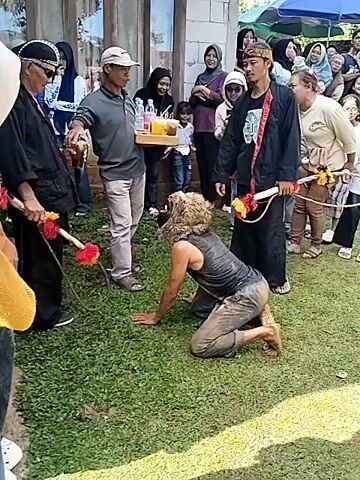 The video shows a traditional ceremony taking place outdoors on a grassy lawn. A person in a lion-like mask is kneeling on all fours, while two men in black outfits stand nearby holding ornate sticks with red and yellow decorations. Another man holds a tray of drinks and offers one to one of the men in black.  A crowd of onlookers, including women in hijabs and children, stands behind them, watching and smiling. A building with a window is visible in the background, along with trees and a blue umbrella. The overall atmosphere suggests a cultural celebration or performance.