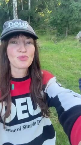 Two women are walking down a dirt path in a wooded area. The woman in the foreground is wearing a black baseball cap, a red, white, and black long-sleeved shirt with text that reads "DEUS House of Simple Pleasures," and she is holding up her left hand in a peace sign gesture. The woman in the background is wearing sunglasses and a gray sweatshirt with a "Hard Rock Cafe" logo. The sun is setting, casting a warm glow on the scene.