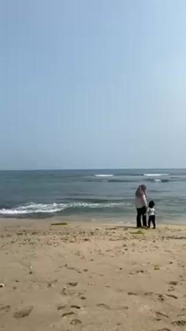 The video shows a beach with a family group. There are three adult women and a small child visible. Two women are standing near the water’s edge, one holding the child’s hand. The other woman is wearing purple clothing. Two other people are crouching down at the water's edge. The beach is sandy, with some dark patches, possibly seaweed or algae. The sea is a dark blue-green with small waves breaking on the shore. The sky is pale blue.