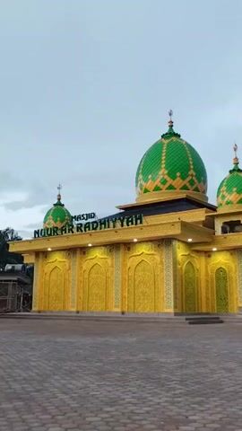 The video features a slow, horizontal pan from left to right, showcasing an exterior view of a mosque. The mosque has bright yellow walls adorned with intricate golden patterns and multiple green domes, also decorated with yellow patterns. The main large dome is centrally located, flanked by smaller domes. On the front facade, the text "MASJID NOOR AR RADHIYYAH" is visible in green lettering. The building is illuminated by yellow lights along its exterior. In the foreground, there is a paved area with a distinctive brick pattern, and a yellow, rectangular pillar or block appears on the right side as the camera pans. The background includes some trees and foliage, with an overcast or twilight sky visible above. No people are visible in the scene.