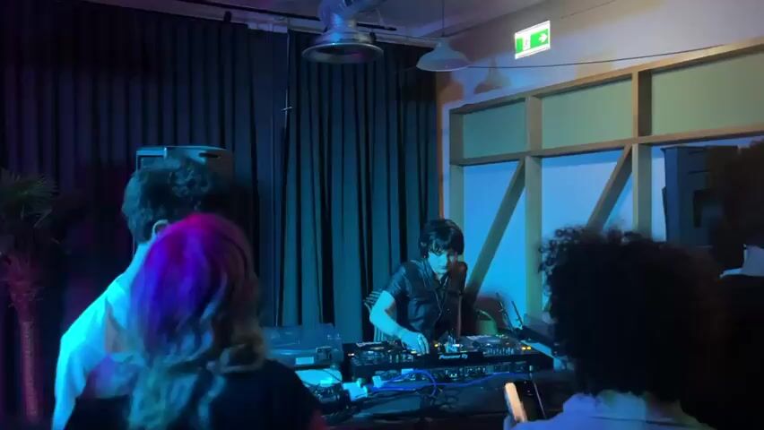 The video shows a DJ, a young person with dark hair and headphones, mixing music at a DJ console in a dimly lit venue with blue lighting. The DJ is wearing a dark short-sleeved leather-like shirt. They are surrounded by people, with the backs of several heads visible. A hand reaches out towards the DJ's equipment. Behind the DJ are dark blue curtains and a geometrically patterned wall. There is sound equipment, including a speaker, visible on the left side of the frame. The atmosphere appears to be that of a party or club.