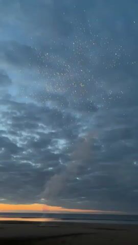A vertical video captures a cloudy sky during what appears to be twilight, with a sliver of orange light visible on the horizon over a body of water. The foreground is dark, suggesting sand or ground. In the sky, there are several small, bright, yellowish lights that could be fireworks or distant lights. A wisp of smoke or cloud is visible rising from the lower part of the sky towards these lights. The clouds are dense and mostly gray, with some lighter patches. The sound in the video includes some indistinct voices, possibly exclamations of awe or surprise, and the faint sound of what could be distant fireworks or a similar explosive event.