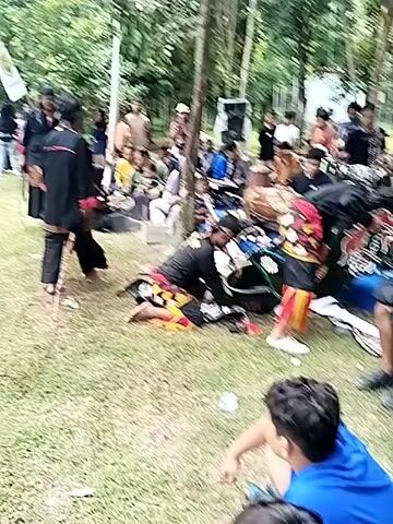 This video captures an outdoor cultural gathering in Puro, Indonesia, focused on what appears to be a traditional Indonesian dance performance or practice, likely Kuda Lumping (Jathilan). Numerous people are present, including performers, musicians, and an audience.

Performers are dressed in traditional attire, featuring black tops, patterned bottoms with red, yellow, and black designs, and some wear elaborate headdresses. One dancer prominently wears a red and yellow skirt-like garment. Several performers are seen on the grassy ground, some appearing to be resting or preparing, while others are standing or moving. Traditional horse props, indicative of Kuda Lumping, are visible, including one with black, green, and red motifs.

Musicians are actively playing instruments, including drums (resembling gamelan percussion) and other percussive elements, with speakers set up nearby. The audience, composed mostly of men in casual attire (t-shirts, hoodies, jeans, shorts), is gathered around the performance area, either sitting on the grass or standing, observing the event. A young boy in a blue shirt is visible in the foreground, facing away from the camera.

The scene is set outdoors on a bright day with green grass and numerous trees in the background, suggesting a park or wooded area. A box labeled "AGUA" is visible on the ground. The atmosphere appears lively and culturally engaged.