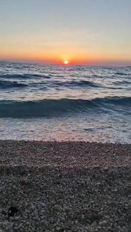 This is a short, vertical video of a beach at sunset. The sun is low on the horizon, casting a warm orange glow across the sky and reflecting on the rippling water. Gentle waves are breaking on a pebbly shore. The scene evokes a sense of peace and tranquility. There is no visible text in the video. The location is identified as Zadar, Croatia. The time of day is sunset. The weather appears to be clear.