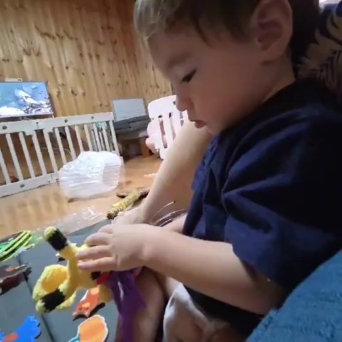 A toddler is seated, wearing a dark blue shirt and diaper. The child is holding a yellow stuffed toy with purple tassels in their hands and bringing it towards their mouth. The toy appears to be a giraffe. The child is on a gray mat that has various other colorful shapes and objects scattered on it, including what looks like pieces of a puzzle or playmat. In the background, there is a wooden paneled wall, a white crib or playpen, and a television screen showing an image of a person. The lighting is warm, suggesting an indoor setting.