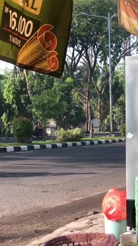 A person wearing a helmet and a black shirt rides a white and green scooter across a paved road. The scooter is moving from left to right in the frame. The road has a black and white striped curb on the left. Behind the curb is a grassy area with trees and bushes. Above the road, a green banner hangs from the left side of the frame, displaying the text "16.000" and "2 WRAPS". A lamppost is visible to the right of the trees. In the foreground, on the right side of the frame, a red plastic bag is placed next to a light-colored object, possibly a bin. A wicker basket is also visible in the lower part of the frame. The scene appears to be outdoors, possibly in a city or suburban area, during daylight.