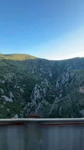 The video captures a scenic view from what appears to be a moving train, looking out over a mountainous landscape near Klopot, Montenegro. The shot begins with blurred metal beams of the train's window or structure quickly passing, then settles on a panoramic view of lush green mountains and deep, rocky valleys under a clear blue sky. The mountainsides are covered with dense vegetation and interspersed with grey, jagged rock formations. The lighting suggests it is daytime, with a soft light illuminating the distant hills. The train continues to move, slowly panning the landscape from left to right, revealing more of the vast natural scenery. No people or visible text are present.