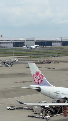 The video shows an airport tarmac on a cloudy day. In the foreground, the tail of a white airplane with a pink flower logo is visible. In the background, another white airplane with blue accents is taking off. Several airport buildings and structures, including hangars and service vehicles, are also present. The focus shifts between the airplanes and the general airport environment.