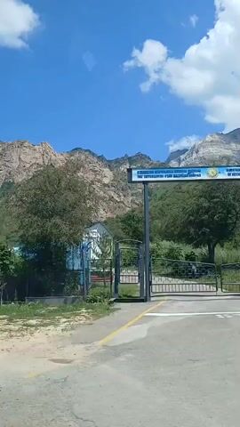 The video shows a scenic view of mountains under a clear blue sky with scattered white clouds. In the foreground, there is a fence with decorative patterns and some greenery. Behind the fence, there are trees and buildings, including what appears to be a hotel or resort complex with a modern architectural style. The camera is moving, suggesting a vehicle is in motion. The overall impression is one of a peaceful, mountainous landscape with some man-made structures integrated into it.