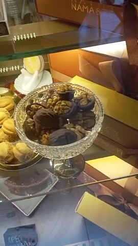This is a close-up, eye-level shot of a display case in a confectionery or bakery. The scene is brightly lit, likely by internal display lights, suggesting it's either daytime or the establishment is open for business. The display case is made of glass, with multiple shelves showcasing various sweets.

On the top shelf, a box with a gold ribbon and the partially visible word "NAMASTE" suggests it might be a gift box or a branded item. Below this, a smaller white label has Russian text that reads "Солона карамель" (Salty Caramel). Next to it is a small, white decorative item resembling a swan or a bird.

The middle shelf is the most populated. To the left, a gold-colored platter holds numerous small, round, golden-brown cookies or pastries, possibly "kurtobey" or similar shortbread. To the right, a clear, ornate glass bowl is filled with what appear to be chocolate-covered walnuts and other nuts. The chocolate-covered walnuts are shaped like actual walnuts.

On the shelf below, a slice of dark chocolate cake or torte is presented on a square white plate. Further down, at the bottom of the frame, there's a glimpse of a dark blue box with white lettering, partially obscured. A visible word on it appears to be "Орео" (Oreo), and another word "ЧИЗКЕЙК" (Cheesecake) is also legible. This indicates the presence of different types of desserts, possibly including cheesecake.

The overall impression is one of a well-stocked and attractive display of desserts, with a focus on pastries, chocolates, and nuts. The items are arranged neatly, suggesting a professional presentation. The presence of Russian text and the location context "Tairove, Ukraine" provide specific clues about the origin of the products and the establishment. No people are visible in the frame, and the activity is static - the display of goods.