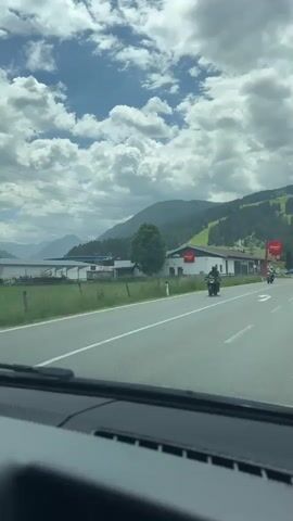 This video appears to be a dashcam recording from a car traveling along a road in a mountainous region, likely Reitdorf, Austria. The scene is set during the daytime under a partly cloudy sky with large, fluffy white clouds.

The primary subjects are vehicles on the road and buildings lining the street. Several motorcycles and cars are visible, including a white Audi station wagon, a dark grey Audi sedan, and a black motorcycle in the foreground. One motorcycle rider is wearing a bright yellow jacket.

The road itself is paved with clear lane markings. Several traffic cones are placed on the road, suggesting a temporary traffic diversion or road work. A distinctive sign for "PENNY MARKT" (Penny Market), a German discount supermarket chain, is prominently displayed on a white building. Another smaller Penny Markt sign is also visible.

The background features rolling green hills and mountains, some of which appear to be ski slopes. A tall electricity pylon is also visible in the distance. The overall atmosphere is of a scenic drive through a semi-rural or suburban area with commercial establishments. There is no discernible emotional tone other than that of a typical journey. The visible text is limited to the "PENNY MARKT" signage.