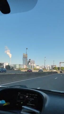 The video shows a view from inside a car driving on a highway. The camera is positioned behind the steering wheel, which is partially visible in the foreground. The road ahead is clear with multiple lanes. Traffic signs are visible overhead, indicating directions to "PARIS-CENTRE" and "A 13 ROUEN" among other destinations. Several cars, including a red bus, are driving on the highway. In the distance, a cityscape with modern buildings and a smokestack is visible under a clear blue sky. The video captures a sunny day.