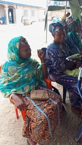 A medium shot shows several seated women in patterned clothing and headwraps at an outdoor event. The woman in the foreground wears a teal headwrap with gold designs and a patterned brown, orange and blue outfit. She wears sandals and has a small, beaded purse on her lap.  She is gesturing with her right hand as if in conversation. To her right, another woman sits in a patterned grey and blue outfit and headwrap. Additional women, partially visible, wear similar attire. The women are seated in simple plastic chairs.  In the background, a building with a white door and window is visible. A tent provides overhead cover.  The ground is dirt.